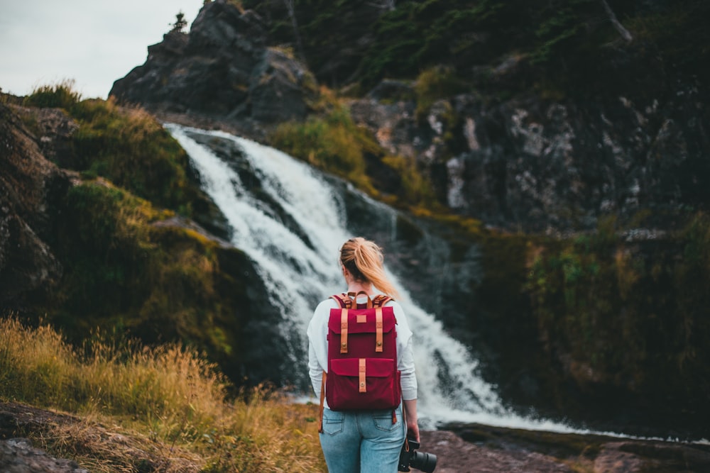 woman in red backpack standing on rocky road looking at waterfalls during daytime