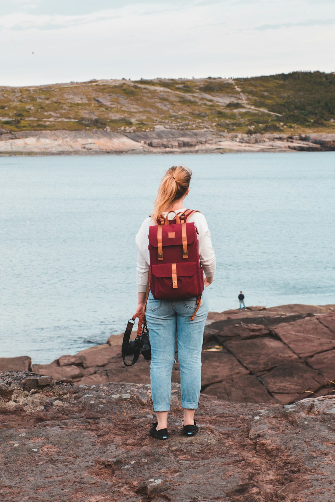 woman in red backpack standing on rock near body of water during daytime