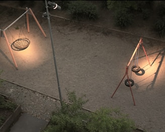 An aerial view of a playground with a gravel surface, highlighting a circular nest swing and a set of swings made from tires, all under warm overhead lighting. The surrounding area includes greenery and shadows cast by the swing set and lighting pole.