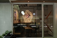 A storefront view featuring a large glass window with a neon sign depicting a cupcake with two cartoon characters inside. Inside, there are tables and chairs, and decorations including wall art and hanging round lights. The name 'Pantry's Best' is displayed on the window.