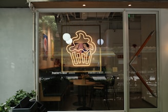 A storefront view featuring a large glass window with a neon sign depicting a cupcake with two cartoon characters inside. Inside, there are tables and chairs, and decorations including wall art and hanging round lights. The name 'Pantry's Best' is displayed on the window.