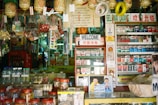 A bustling traditional store is filled with colorful packaged goods, hanging decorations, and an array of tobacco products on display. A prominent 'Welcome' sign in multiple languages greets visitors amidst an eclectic mix of snacks, bottled water, and various items. Brightly wrapped candies and snacks hang from the ceiling, adding to the vibrant atmosphere.