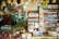 A bustling traditional store is filled with colorful packaged goods, hanging decorations, and an array of tobacco products on display. A prominent 'Welcome' sign in multiple languages greets visitors amidst an eclectic mix of snacks, bottled water, and various items. Brightly wrapped candies and snacks hang from the ceiling, adding to the vibrant atmosphere.