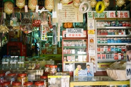 A bustling traditional store is filled with colorful packaged goods, hanging decorations, and an array of tobacco products on display. A prominent 'Welcome' sign in multiple languages greets visitors amidst an eclectic mix of snacks, bottled water, and various items. Brightly wrapped candies and snacks hang from the ceiling, adding to the vibrant atmosphere.