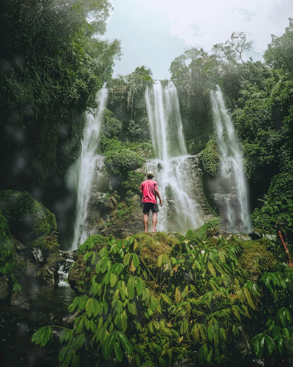 man in red shirt standing on rock near waterfalls during daytime photo –  Free Bali Image on Unsplash