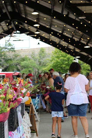 A bustling farmers market with people walking under strings of lights. Stalls are lined with colorful bouquets of flowers, and a diverse group of people mingle, including a child holding a bouquet. The market is sheltered under a large canopy, and trees are visible in the background.