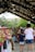 A bustling farmers market with people walking under strings of lights. Stalls are lined with colorful bouquets of flowers, and a diverse group of people mingle, including a child holding a bouquet. The market is sheltered under a large canopy, and trees are visible in the background.