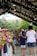 A bustling farmers market with people walking under strings of lights. Stalls are lined with colorful bouquets of flowers, and a diverse group of people mingle, including a child holding a bouquet. The market is sheltered under a large canopy, and trees are visible in the background.