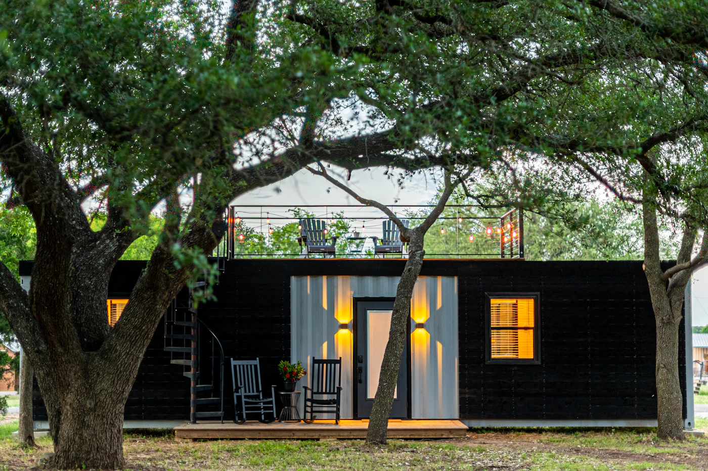 Black-clad container home with porch chairs and warm lit windows under a tree