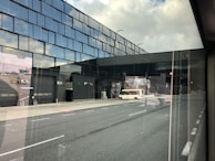 A modern airport terminal with reflective glass windows, showcasing a runway area outside. There is a white vehicle parked near the entrance labeled R24, and various airport equipment can be seen in the background.