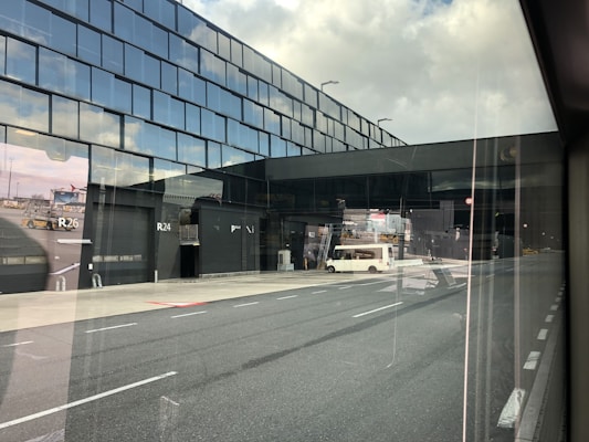 A modern airport terminal with reflective glass windows, showcasing a runway area outside. There is a white vehicle parked near the entrance labeled R24, and various airport equipment can be seen in the background.