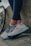 A pair of legs in dark leggings are leaning against a concrete surface next to a bicycle wheel. The person is wearing light-colored athletic shoes with intricate patterns and details, featuring shades of gray, blue, and red.