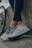 A pair of legs in dark leggings are leaning against a concrete surface next to a bicycle wheel. The person is wearing light-colored athletic shoes with intricate patterns and details, featuring shades of gray, blue, and red.
