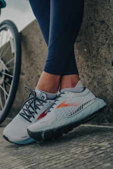 A pair of legs in dark leggings are leaning against a concrete surface next to a bicycle wheel. The person is wearing light-colored athletic shoes with intricate patterns and details, featuring shades of gray, blue, and red.