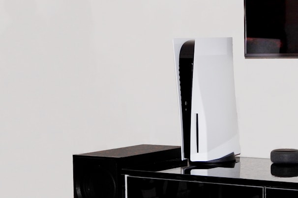 A white, modern gaming console stands vertically on a black surface next to a black speaker. In the background, part of a flat-screen television is visible on the wall.