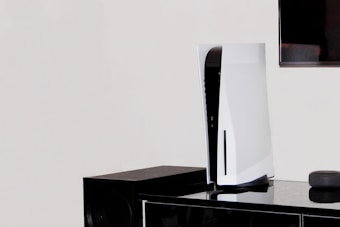 A white, modern gaming console stands vertically on a black surface next to a black speaker. In the background, part of a flat-screen television is visible on the wall.