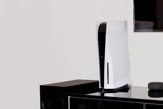 A white, modern gaming console stands vertically on a black surface next to a black speaker. In the background, part of a flat-screen television is visible on the wall.