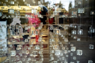 Numerous transparent cubes are suspended in rows, creating a layered effect with reflections and depth. In the background, blurred hints of pink flowers and interior elements suggest a display setting.