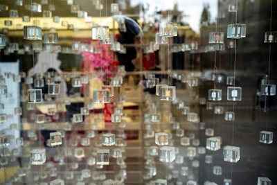 Numerous transparent cubes are suspended in rows, creating a layered effect with reflections and depth. In the background, blurred hints of pink flowers and interior elements suggest a display setting.