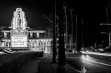 A black and white photograph featuring a classic cinema marquee with neon lights displaying the names of movies. The street beside the cinema is illuminated by streaks of light from passing cars, and tall palm trees can be seen in the background. The atmosphere suggests a quiet, nighttime urban environment.