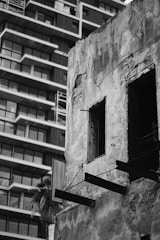 A contrast between an old, deteriorated concrete building and a modern apartment complex with a grid of balconies is evident. The old building shows significant wear with exposed bricks and missing windows, indicating abandonment or decay.