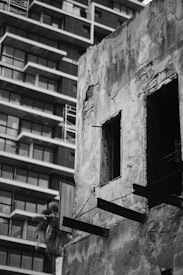 A contrast between an old, deteriorated concrete building and a modern apartment complex with a grid of balconies is evident. The old building shows significant wear with exposed bricks and missing windows, indicating abandonment or decay.