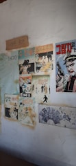 The image depicts a wall decorated with vintage comic book posters and a calendar from the year 1970. The posters feature various illustrated characters, predominantly in Korean text, and include elements such as a man holding a gun, a woman, and other stylized figures. There is also a piece of paper with handwritten text.