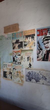 The image depicts a wall decorated with vintage comic book posters and a calendar from the year 1970. The posters feature various illustrated characters, predominantly in Korean text, and include elements such as a man holding a gun, a woman, and other stylized figures. There is also a piece of paper with handwritten text.