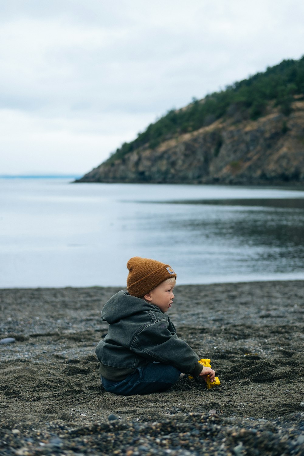 Person in grauem Kapuzenpullover und brauner Strickmütze sitzt auf grauem Felsen in der Nähe von Wasser