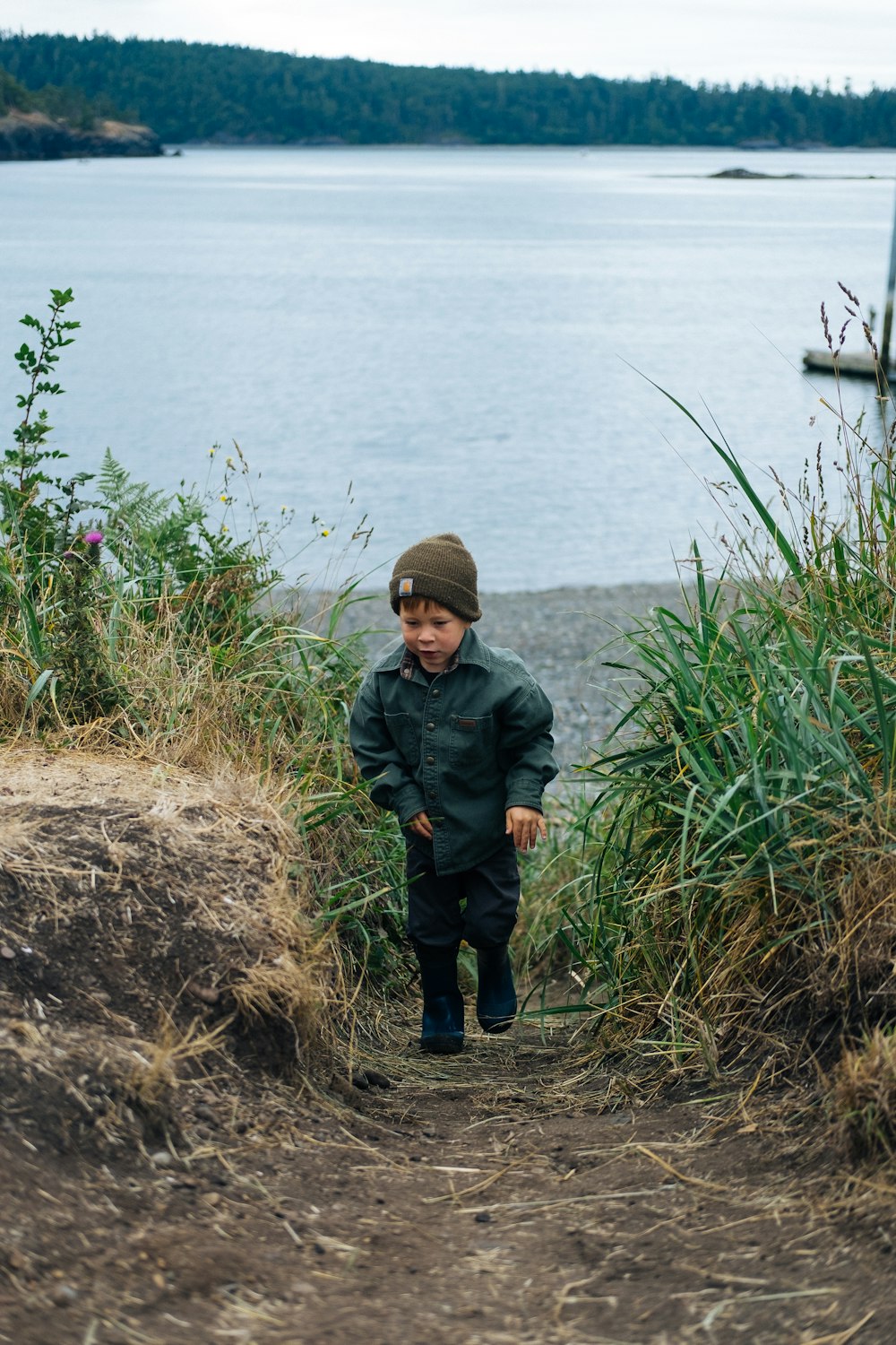 黒いジャケットと黒いズボンを着た男の子が、水域の近くの茶色の草の上に立っています