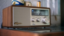 A retro-style radio with a wooden exterior and a metallic speaker on the left side. The control panel includes dials for volume, frequency, and tuning. The frequency display shows AM and FM bands with blue backlighting. Objects in the background include a wall and small items, such as batteries.