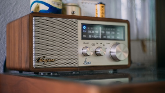 A retro-style radio with a wooden exterior and a metallic speaker on the left side. The control panel includes dials for volume, frequency, and tuning. The frequency display shows AM and FM bands with blue backlighting. Objects in the background include a wall and small items, such as batteries.