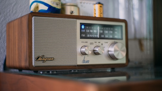 A retro-style radio with a wooden exterior and a metallic speaker on the left side. The control panel includes dials for volume, frequency, and tuning. The frequency display shows AM and FM bands with blue backlighting. Objects in the background include a wall and small items, such as batteries.