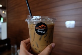 A hand holding a plastic cup filled with iced coffee, featuring a black straw. The cup has a logo with text in a stylish font, and the background consists of wooden paneling, creating a cozy caf&eacute; atmosphere.