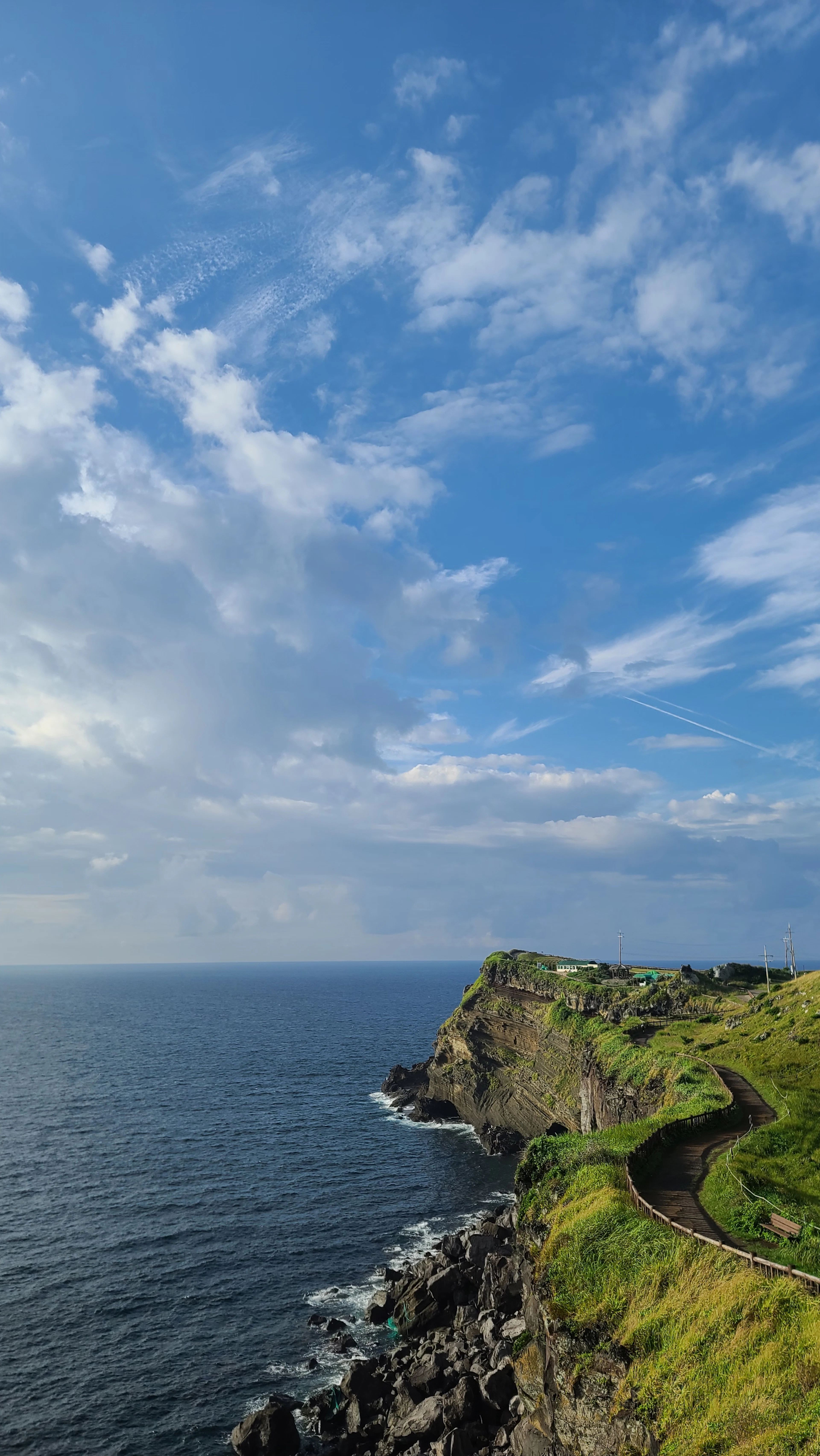 A grassy coastal walking path winding along dark volcanic sea cliffs on Jeju Island under a wide blue sky