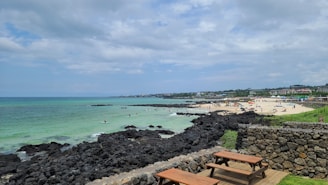 A scenic coastal area features a sandy beach populated with people enjoying various activities under colorful umbrellas. The ocean water appears clear with shades of blue and green, and there are dark volcanic rocks along the shoreline. The weather is partly cloudy with a mix of sun and clouds. Nearby, a stone wall encloses a grassy area where wooden picnic tables are set up, offering an inviting place to relax and enjoy the view.