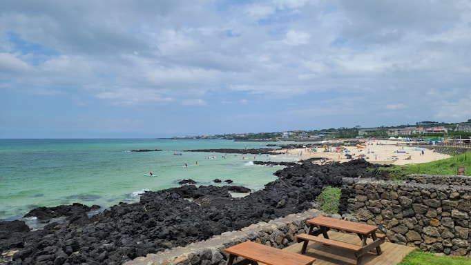 A scenic coastal area features a sandy beach populated with people enjoying various activities under colorful umbrellas. The ocean water appears clear with shades of blue and green, and there are dark volcanic rocks along the shoreline. The weather is partly cloudy with a mix of sun and clouds. Nearby, a stone wall encloses a grassy area where wooden picnic tables are set up, offering an inviting place to relax and enjoy the view.