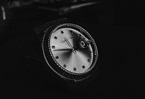 A close-up view of a luxury watch with a shiny, metallic face. The watch features a smooth bezel with an engraved design, metallic hour markers, and distinct watch hands. The brand name is visible near the top of the watch face. The image is in black and white, highlighting the reflective surface and intricate details of the watch.
