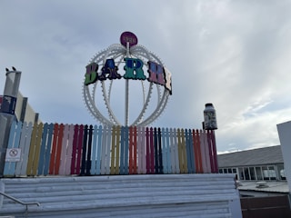 A multicolored picket fence is positioned in front of a large, decorative sign that reads 'BABY' with an establishment date of 1926 on top. The sign structure is spherical with lights, and a large jar filled with colorful items is on one side. The sky is partly cloudy, creating a bright and partially shadowed ambiance.