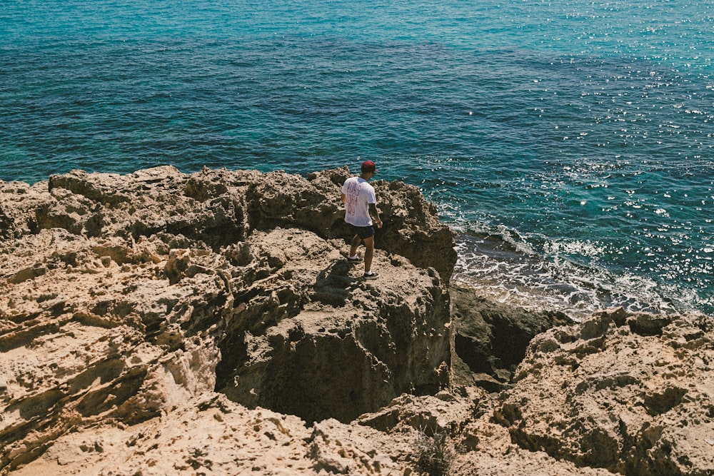 man in white t-shirt standing on rock formation near body of water during daytime