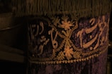 A richly decorated fabric featuring intricate gold embroidery on a deep purple background. The design includes floral patterns and elegant script, with a fringe trimming the top edge.