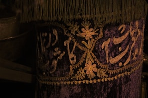 A richly decorated fabric featuring intricate gold embroidery on a deep purple background. The design includes floral patterns and elegant script, with a fringe trimming the top edge.