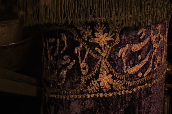 A richly decorated fabric featuring intricate gold embroidery on a deep purple background. The design includes floral patterns and elegant script, with a fringe trimming the top edge.