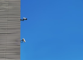 A modern building facade with horizontal metal slats is juxtaposed against a clear blue sky. Two surveillance cameras are mounted at different heights on the wall, both facing outward.