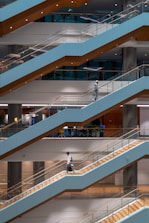 A multi-level modern interior with parallel escalators running diagonally across the space. Two individuals are walking on separate levels, one carrying a bag and the other in a suit. The area is well-lit with warm lighting and visible office spaces behind glass panels.