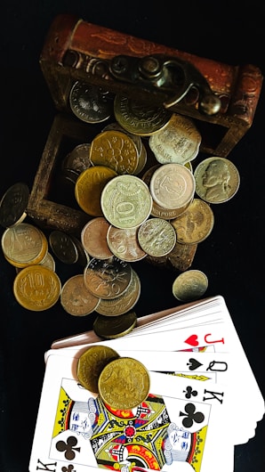 An open wooden treasure chest overflowing with various coins, both silver and gold in color, is paired with a deck of playing cards that are spread out next to it. The cards feature the Jack and King of clubs and hearts, prominently displayed.