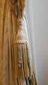 An elegant curtain with intricate tassel tiebacks featuring braided and twisted cords. The fabric has a subtle floral pattern and a smooth texture, draped gracefully to one side.