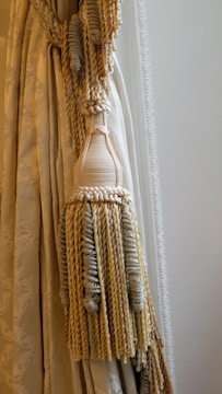 An elegant curtain with intricate tassel tiebacks featuring braided and twisted cords. The fabric has a subtle floral pattern and a smooth texture, draped gracefully to one side.