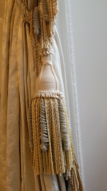 An elegant curtain with intricate tassel tiebacks featuring braided and twisted cords. The fabric has a subtle floral pattern and a smooth texture, draped gracefully to one side.