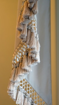A close-up view of a textile with intricate macramé patterns and numerous fringed tassels in neutral tones. The woven design features a combination of tightly wrapped and loosely knotted textures, creating a sense of handcrafted elegance.